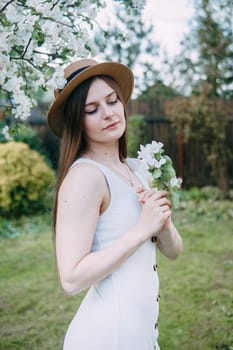 Beautiful young girl in white dress and hat in blooming Apple orchard. Blooming Apple trees with white flowers