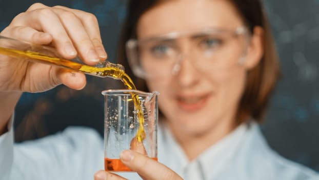 Closeup of young beautiful teacher focus on mixing chemical liquid at laboratory. Skilled scientist doing an experiment by inspect colored solution while standing in front of blackboard. Erudition.