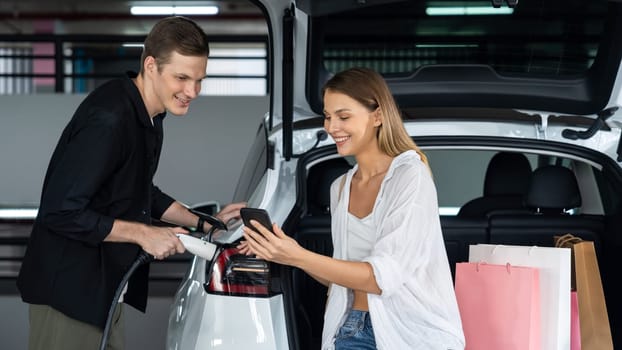 Young couple travel with EV electric car to shopping center parking lot charging in downtown city showing urban sustainability lifestyle by green clean rechargeable energy of electric vehicle innards