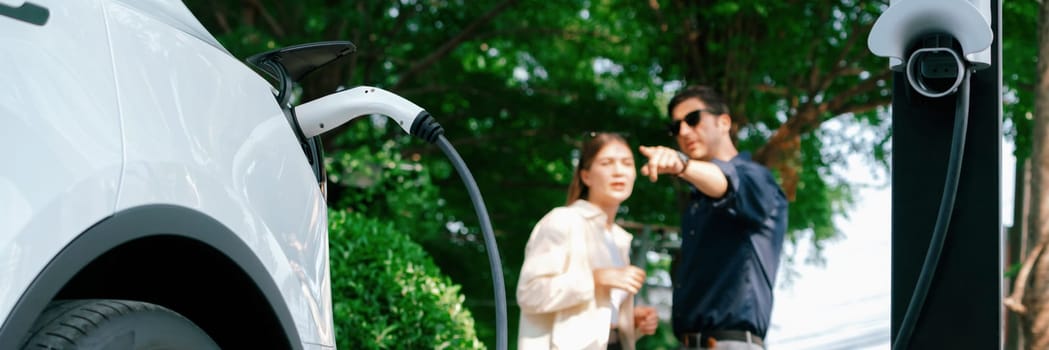 Young couple recharge electric car battery from charging station in green city park in springtime. Rechargeable EV car for sustainable environmental friendly urban travel lifestyle. Panorama Expedient