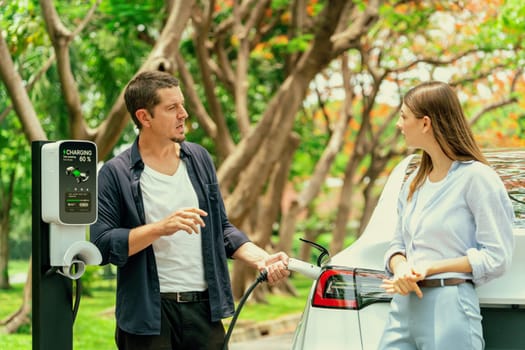 Lovely young couple recharging battery for electric car during autumnal road trip travel EV car in autumnal forest. Eco friendly travel on vacation during autumn. Exalt