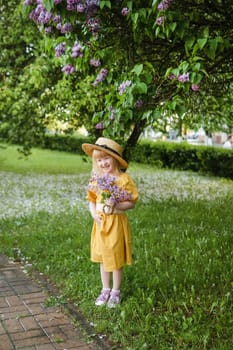 A little girl in a yellow dress and straw hat wearing a bouquet of lilacs. A walk in a spring park, blossoming lilacs