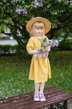 A little girl in a yellow dress and straw hat wearing a bouquet of lilacs. A walk in a spring park, blossoming lilacs