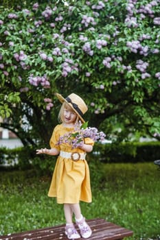 A little girl in a yellow dress and straw hat wearing a bouquet of lilacs. A walk in a spring park, blossoming lilacs