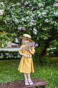 A little girl in a yellow dress and straw hat wearing a bouquet of lilacs. A walk in a spring park, blossoming lilacs