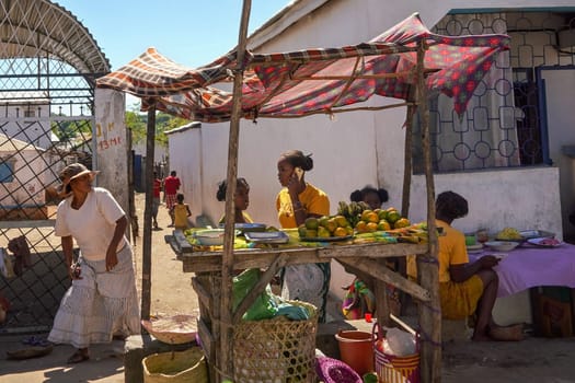 Sakaraha, Madagascar - May 05, 2019: Unknown Malagasy women selling fruits by the main road at simple wooden stall. There are not many shops on Madagascar and food is usually sold on streets.
