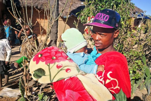Ilakaka, Madagascar - April 30, 2019: Unknown Malagasy man in fitted cap holds his baby on hands next to road on sunny day, simple clay house in background. People of Madagascar are poor but cheerful
