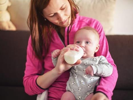 Young mother holding her infant baby son on her hands, feeding him milk from bottle