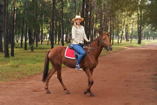 Young woman in shirt and straw hat, riding brown horse, blurred background with trees in park