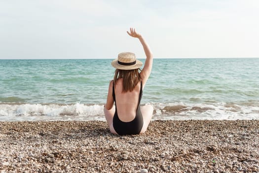 Woman in black swimsuit and straw hat on beach, seaside. View from the back. Summer vacation at the sea