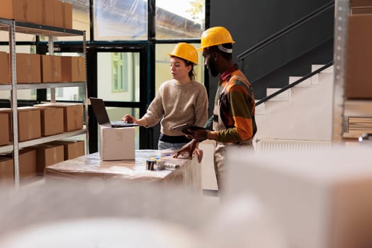 Warehouse supervisors checking online orders on laptop computer, analyzing shipping details while preparing packages. African american worker analyzing merchandise logistics in storage room