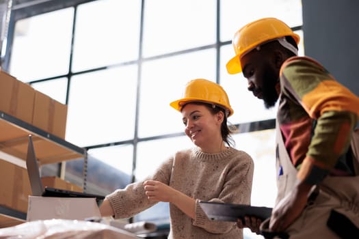 Diverse coworkers wearing industrail overall and helmet, while checking online orders on laptop computer in storage room. Stockroom team analyzing delivery details on digital device in warehouse