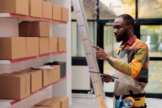 African american worker in industrial overall checking boxes, working at customers order preparing products for delivery in storehouse. Employee managing parcels transportation in warehouse