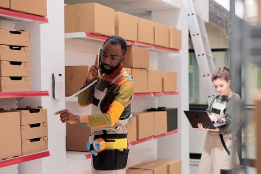 African american manager discussing merchandise logistics with remote supervisor at landline phone in warehouse. Employee working at customers orders, checking shipment details in storehouse
