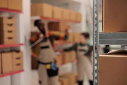 Selective focus of metallic shelves full with cardboard boxes, diverse employee working at customers orders delivery in warehouse. Worker discussing goods logistics with manager at landline phone