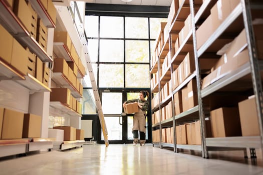 Supervisor woman carrying cardboard box during inventory, listening music at headphone in warehouse. Storage room manager preparing customers orders before shipping products in storehouse
