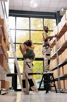 Storehouse workers looking at shelves full with clients orders, carrying cardboard box while listening music at headphones. Employees working at products packages preparing delivery in warehouse