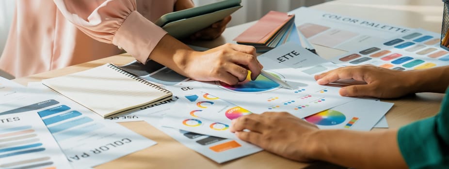 Skilled architect searching data from tablet while selecting an appropriate color from color wheel at table with color palette and document scatter around. Creative design concept. Variegated.