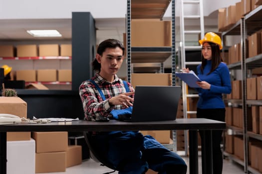 Young asian delivery service worker monitoring parcel transportation on laptop. Freight supply chain logistics manager working on computer while sitting at industrial warehouse desk