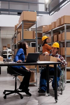 Delivery manager and coworker in wheelchair preparing parcels for shipping while managing orders from laptop at a warehouse desk. Storehouse worker with disability giving packed box to colleague