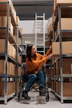 Storehouse asian order picker in wheelchair taking parcel from rack to pack product before dispatching. Retail business warehouse young woman package handler choosing box on shelf