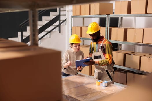 Diverse supervisors looking at merchandise logistics report, while preparing customers orders in storehouse. African american employee working at products inventory with manager in storage room