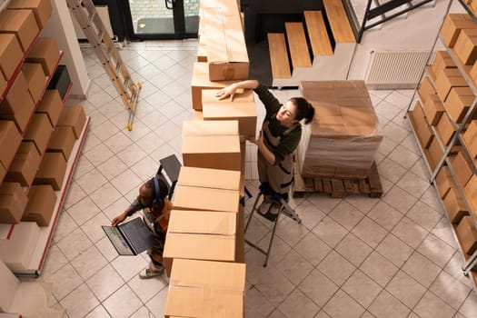 Top view of multi ethnic colleagues working in storehouse, preparing customers orders before start delivery merchandise. Storage room team in protective overall working with cardboard boxes