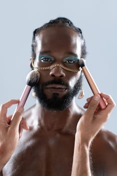 Portrait of African American gay man applying makeup with brushes held by crop artist. Closeup of homosexual man wearing bright eyeshadow and gold chain looking at camera on blue background.