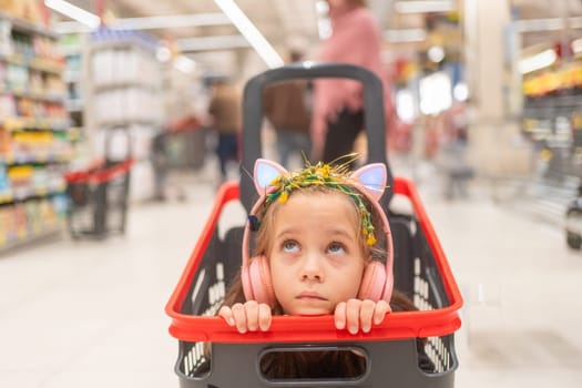 Thoughtful girl listening to music through wireless headphones. Female child looking up while sitting in shopping cart at grocery store.