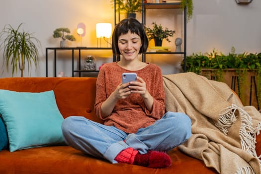 Happy young woman in casual clothes thinking and text messaging on smartphone. Hispanic girl sitting on sofa and using mobile phone in living room at home. Lady watching online movie in apartment.