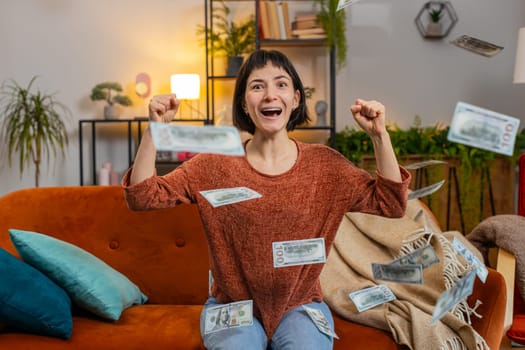 Happy joyful Caucasian woman fingers crossed shouting, celebrating success, winning, lottery jackpot goal achievement, good news victory. Money cash dollar rain falling on smiling girl on sofa at home