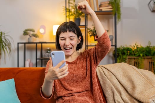 Surprised shocked woman browsing smartphone winning lottery sitting on sofa in home room. Excited Caucasian girl celebrating success victory clenching fists in apartment. Happy lady play online game
