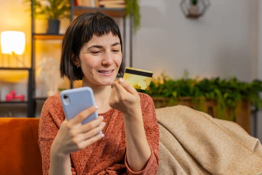 Portrait of happy woman make payments online purchase shopping internet store. Smiling Caucasian girl holds banking credit card involved in verification process in mobile application on sofa at home
