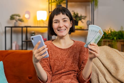 Smiling happy woman counting money dollar banknotes using smartphone calculator on sofa in room at home. Hispanic lovely girl calculating budget, savings, earnings and planning vacation in apartment