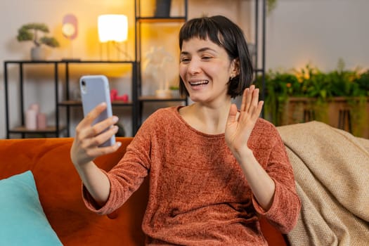 Smiling young woman in casual clothes waving on video call via smartphone in apartment. Happy Caucasian girl having conversation with friend on mobile phone sitting on sofa in living room at home.