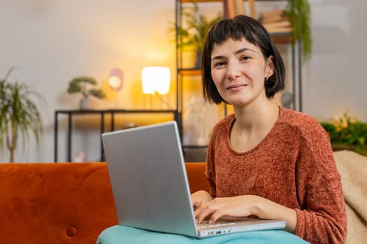 Young student girl sitting on sofa finishing laptop work study education project at home room. Smiling happy woman freelancer working online, remote job sitting on sofa looking at camera in apartment.