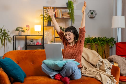 Surprised young woman rising hands using laptop receive good news shocked by sudden victory celebrate lottery jackpot win in apartment. Cheerful girl with laptop sits on couch in living room at home