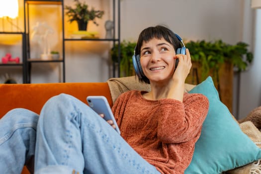 Happy Caucasian girl in wireless headphones choosing listening favorite energetic dancing music on smartphone. Smiling young woman relaxing on sofa in living room at home in apartment during weekend.