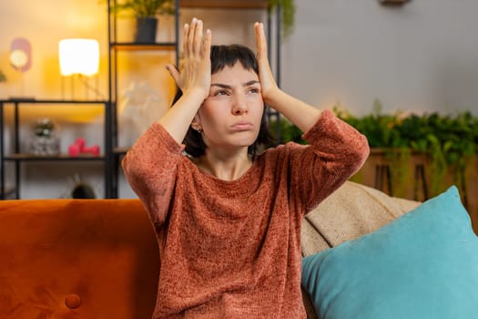 Displeased Caucasian young woman rubbing temples to cure headache problem, suffering from tension and migraine, stress grimacing in pain. Upset girl sitting on couch in living room at home apartment
