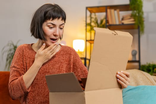 Happy Hispanic young woman unpacking delivery parcel sitting at home. Smiling satisfied girl shopper, online shop customer opening cardboard box receiving purchase gift by fast postal shipping