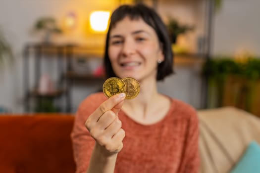 Portrait of smiling Caucasian woman holding two golden bitcoins BTC, looking at camera. Happy surprised girl stock trader showing thumbs up sitting on sofa in living room at home. Increasing wealth.