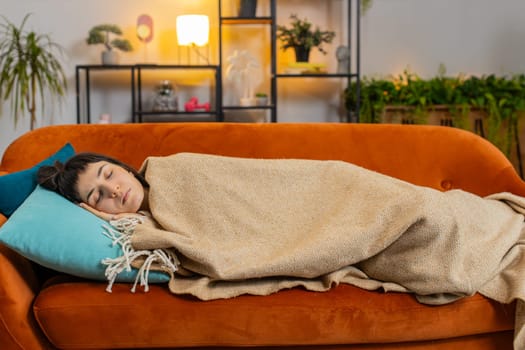 Tired Caucasian woman sleeping quietly covering with blanket on sofa. Happy girl smiling lying on couch sleeping on comfortable on cushion resting peacefully in living room. Healthy sleep concept.
