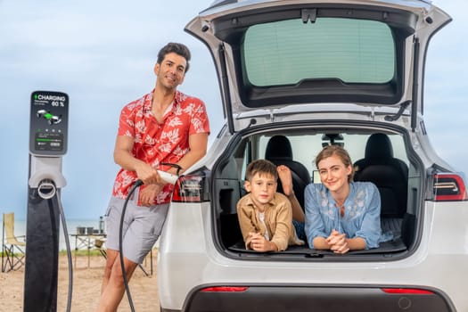 Family vacation trip traveling by the beach with electric car, lovely family sit on the trunk, charging EV car battery with green and sustainable energy. Family travel and eco-friendly car. Perpetual