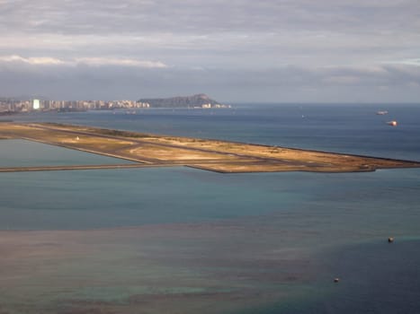 Stunning bird’s-eye view of a long and narrow runway in Honolulu, Hawaii, surrounded by the blue ocean and the city skyline. The image showcases the beauty and uniqueness of the island’s airport, which is one of the largest and busiest in the United States.