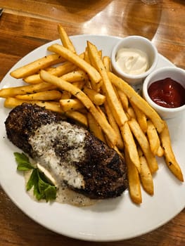 A succulent grilled steak topped with a creamy sauce, accompanied by a generous serving of golden crispy fries, presented on a white plate. The perfect image to represent a hearty, delicious meal.