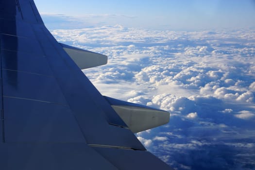 View from an airplane window, focusing on the aircraft’s wing that is visible against a backdrop of clouds and sky. The photo shows the grey wing with smooth surfaces and streamlined design, and the vast expanse of fluffy white clouds scattered across the blue sky.