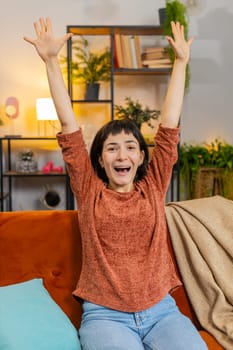 Portrait of happy woman shouting, celebrating success victory, winning, watching TV championship clapping. Cheerful girl rising hands having fun sitting on couch in living room at home. Vertical shot