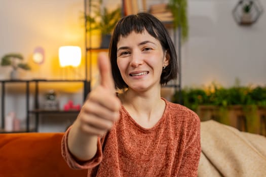 Portrait of excited Caucasian woman looking approvingly at camera showing double thumbs up, like sign positive, good news, positive feedback. Smiling girl sitting on couch in living room at home.