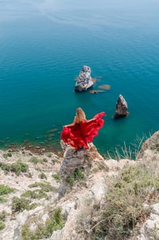 A girl with loose hair in a long red dress descends the stairs between the yellow rocks overlooking the sea. A rock can be seen in the sea. Sunny path on the sea from the rising sun.