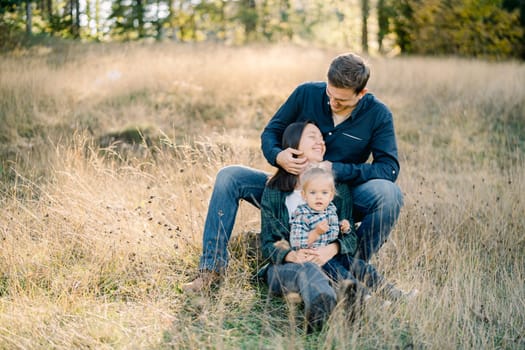 Dad stroking mom hair with a little girl on her lap sitting behind her on the lawn. High quality photo